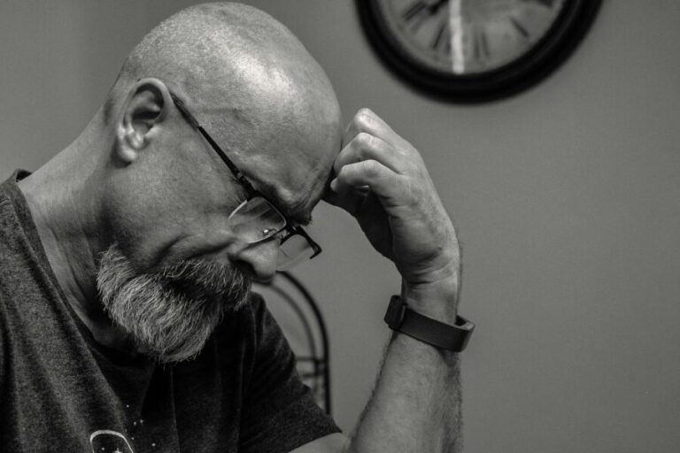 Black and white portrait of a thoughtful bald man indoors, capturing a moment of reflection with a wall clock in the background.